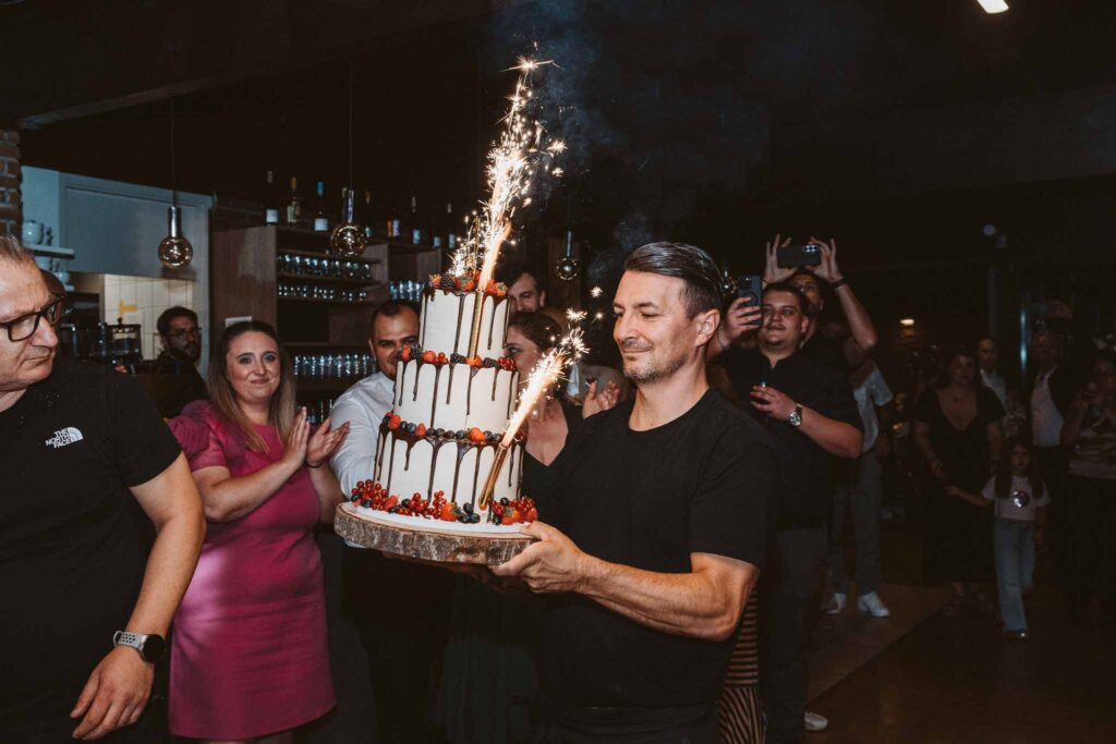 Eine Hochzeitstorte mit brennenden Wunderkerzen wird feierlich in die Partylocation in Böblingen getragen, umgeben von applaudierenden Gästen.