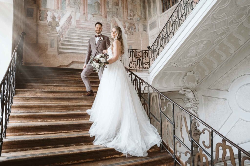 Paar steht auf der Holztreppe im Residenzschloss Ludwigsburg, während warmes Sonnenlicht durch das Fenster einfällt.