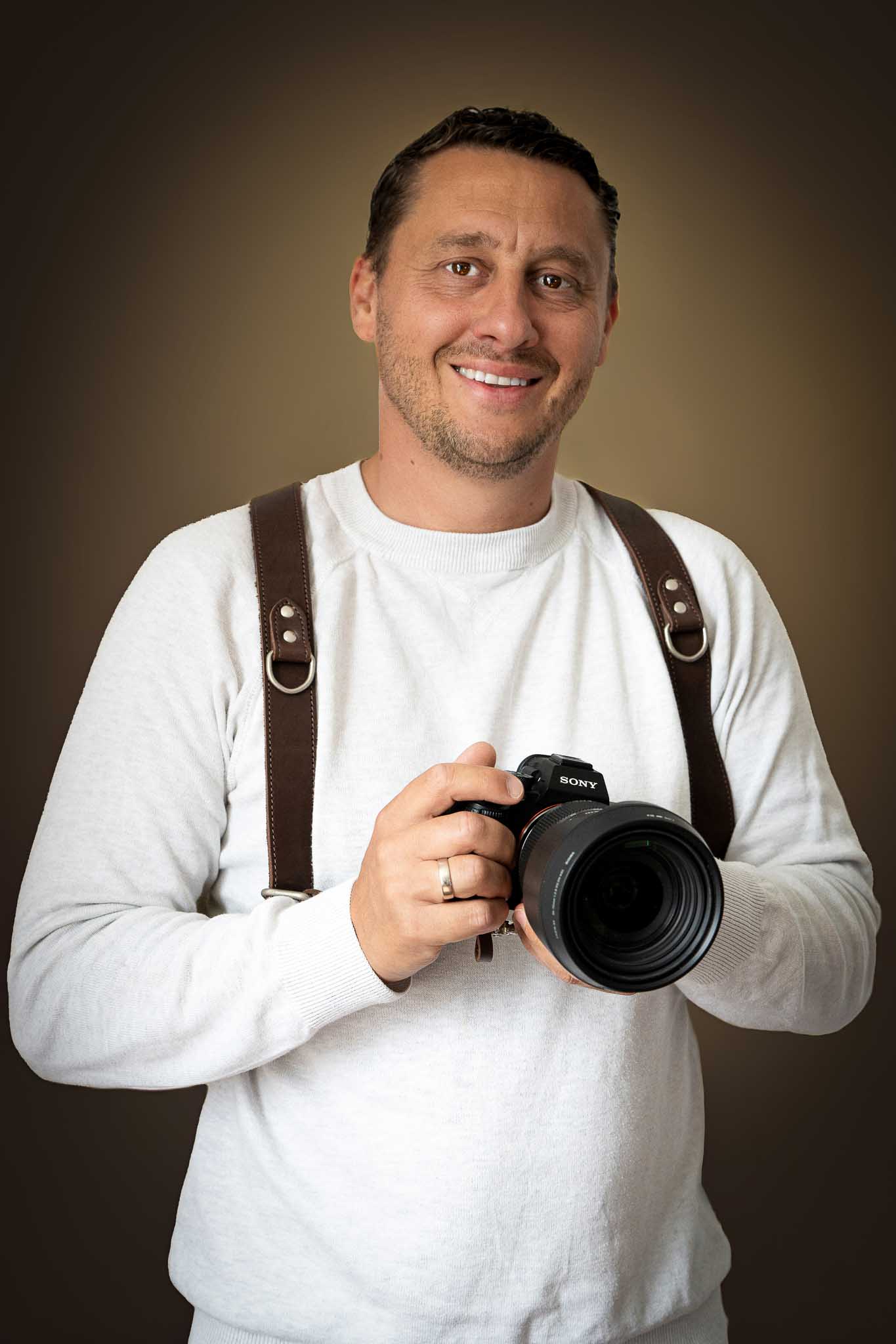 Business-Portrait eines Fotografen mit Kamera und Tragegurt vor neutralem Hintergrund.