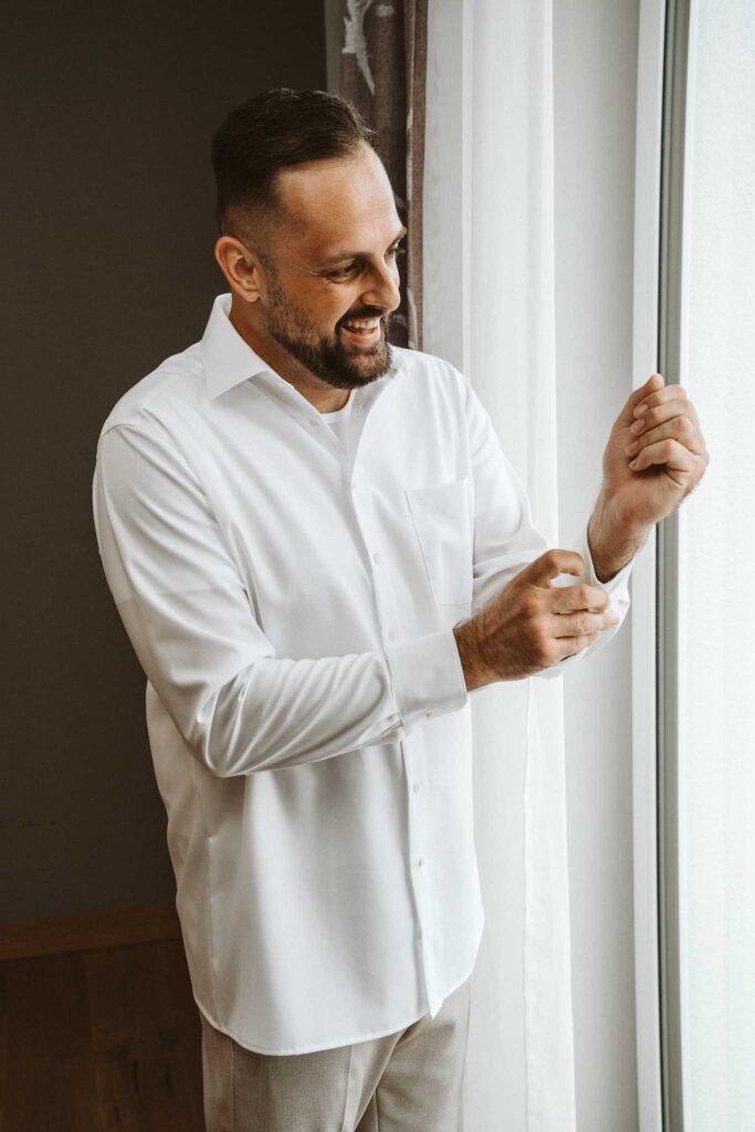 Bräutigam lacht am Fenster, während er seinen Hemdsärmel hochkrempelt – Getting Ready in Breisach am Rhein.