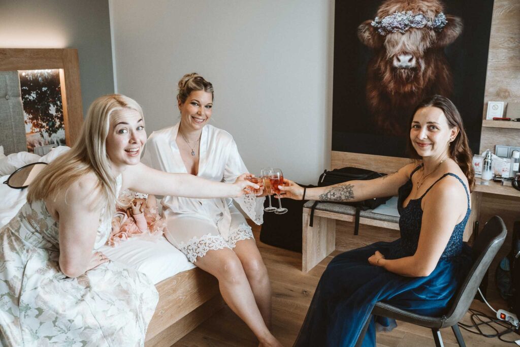 Drei Frauen beim entspannten Getting Ready im Hotel, stoßen gemeinsam mit Sekt an.