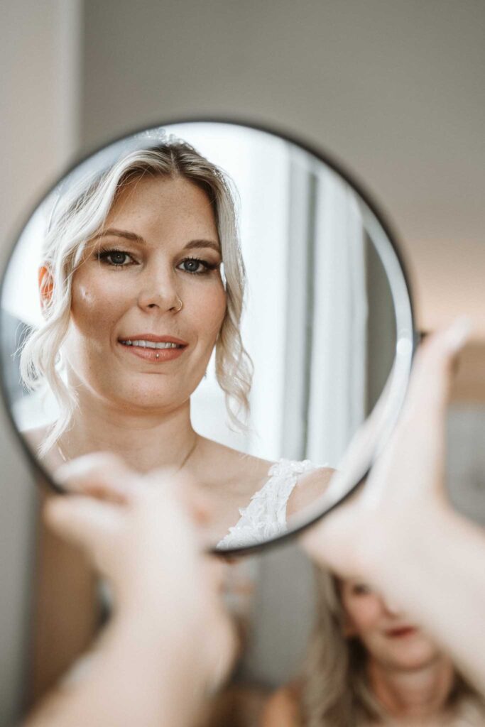 Braut betrachtet sich lächelnd im Spiegel während des Getting Ready am Hochzeitstag in Breisach am Rhein.