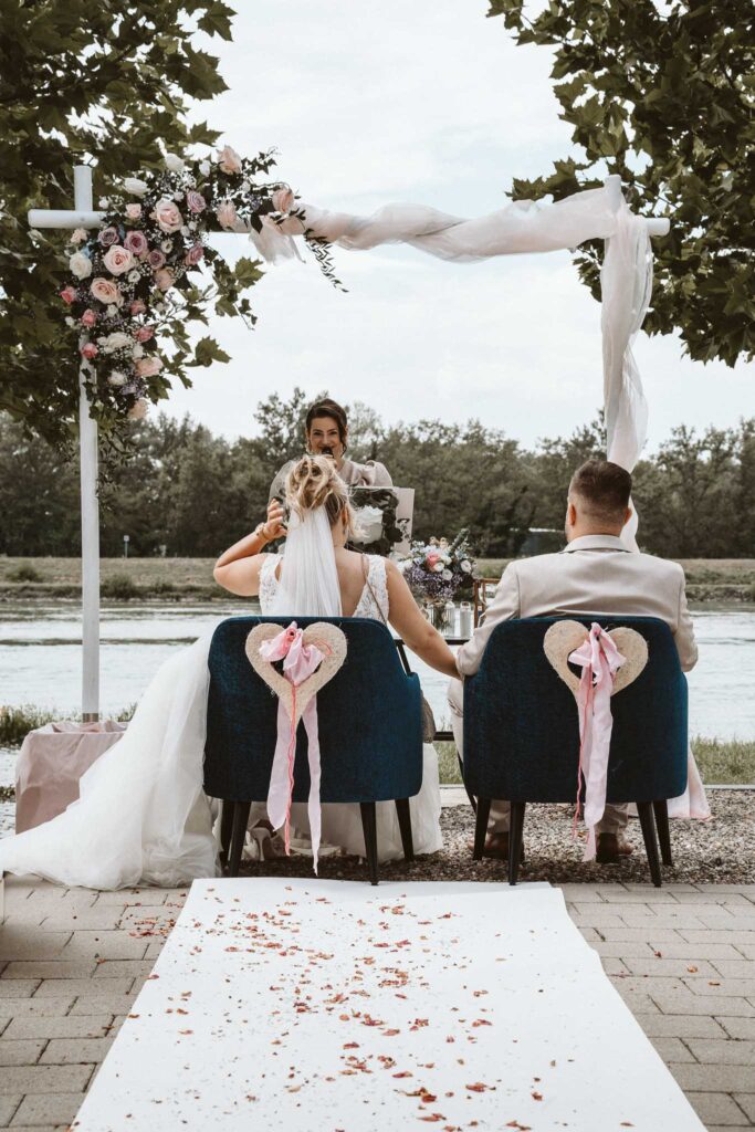 Freie Trauung in Breisach am Rhein – Braut und Bräutigam sitzen Hand in Hand vor einem floralen Traubogen direkt am Wasser.