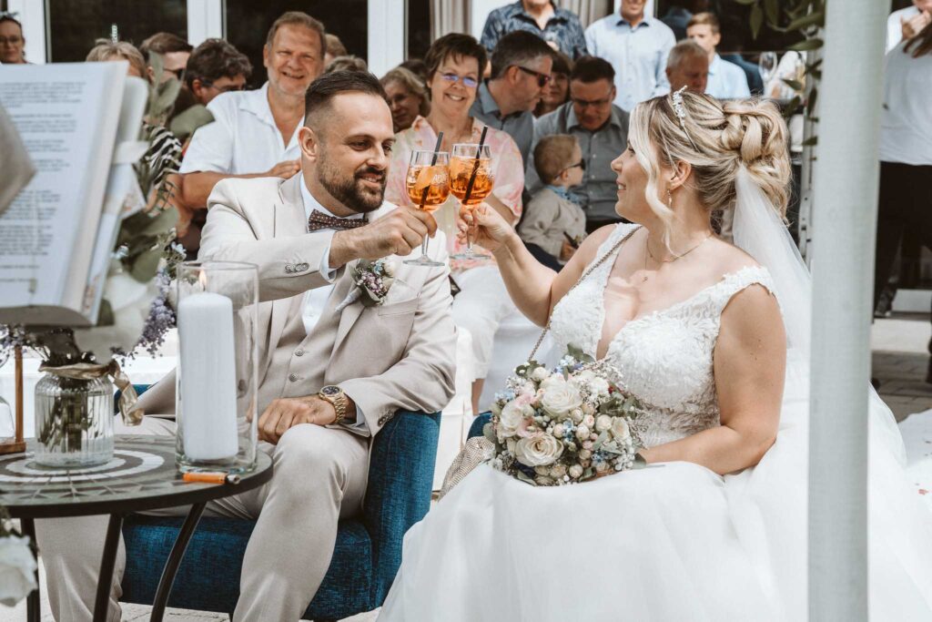 Brautpaar stößt nach der Trauung in Breisach am Rhein mit Aperol an, umgeben von Gästen, während sie lachend auf ihrer Hochzeitsfeier anstoßen.