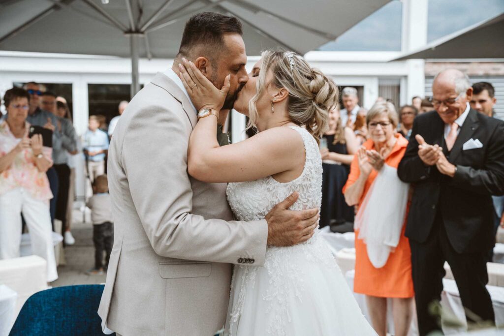 Braut und Bräutigam küssen sich nach der Trauung in Breisach am Rhein, umgeben von applaudierenden Gästen im Außenbereich.