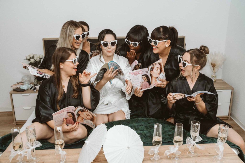 Braut und ihre Freundinnen sitzen gemeinsam beim Getting Ready und stöbern lachend in Hochzeitsmagazinen.