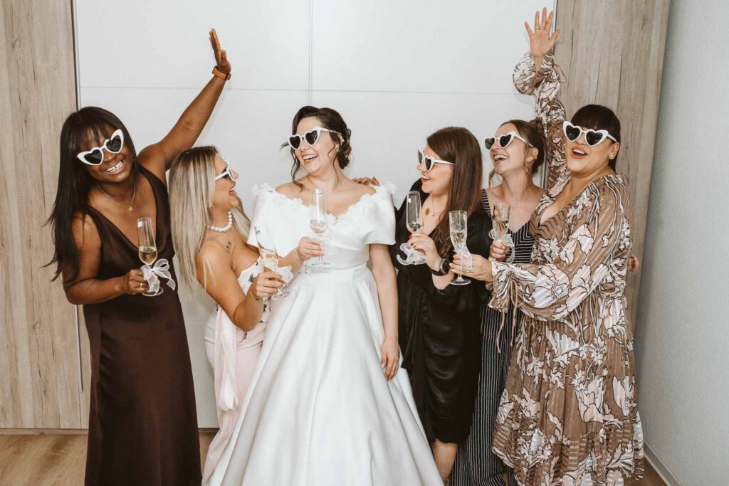 Braut und ihre engsten Freundinnen feiern gemeinsam beim Getting Ready, alle tragen Sonnenbrillen und lachen ausgelassen.