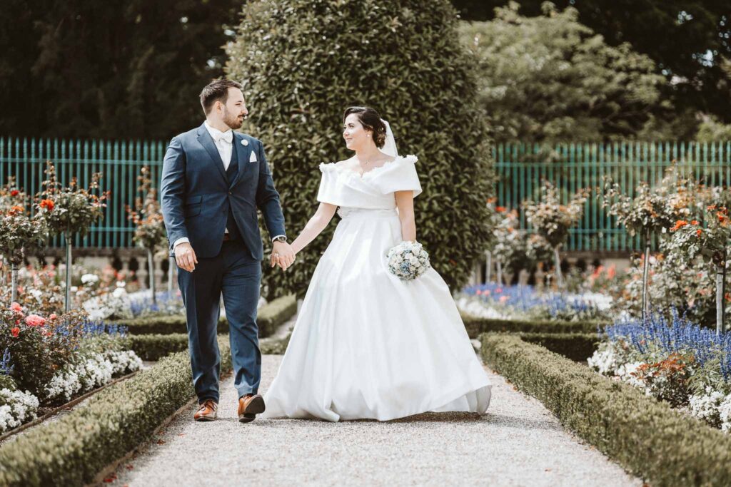 Brautpaar schlendert Hand in Hand durch den Burggarten des Barockschlosses Ludwigsburg und lächelt sich glücklich an.