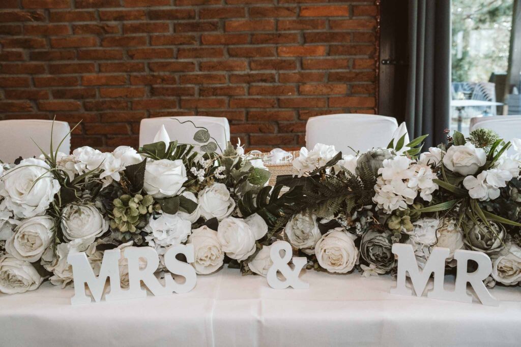 Elegant dekorierte Hochzeitstafel mit weißer Blumenarrangement-Girlande und Mr & Mrs Schriftzug in moderner Location.